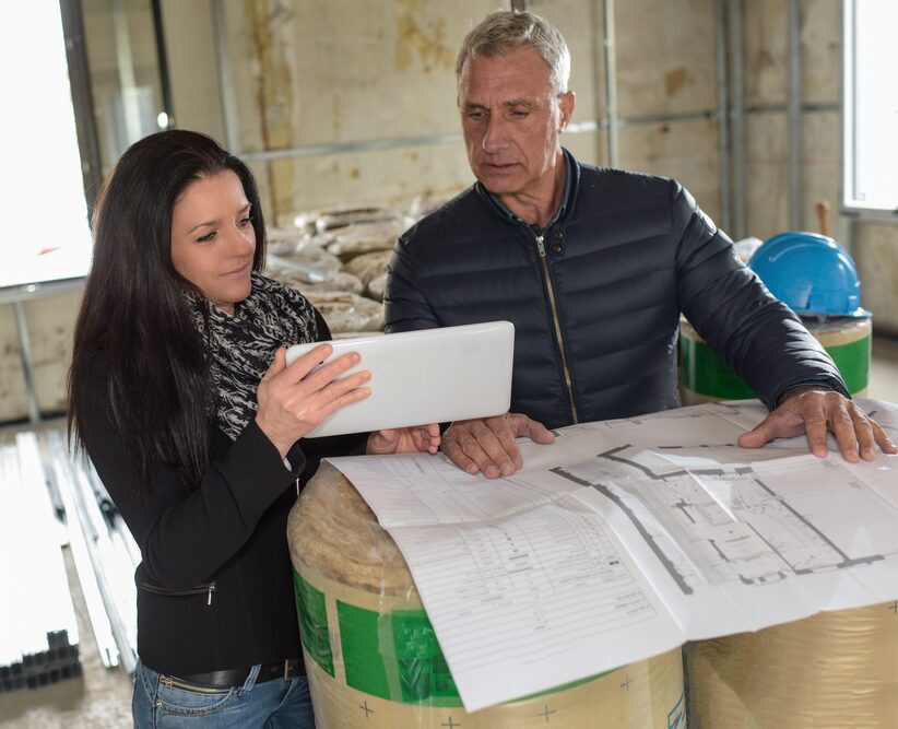A man and a woman standing inside an unfinished building site, reviewing architectural blueprints and referencing a tablet.